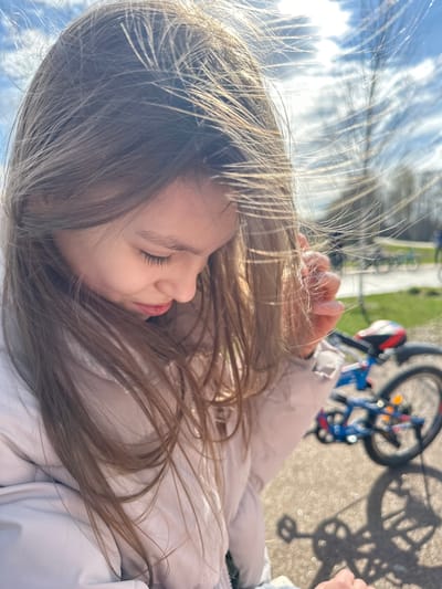 A close-up, tilted perspective shows a young girl with long brown hair lying down. Her eyes are closed, and she appears to be resting. A hand is visible near her head. In the blurred background, a bicycle, a paved path, and some greenery are discernible. The location is likely in a park area in Fryazino, Russia.