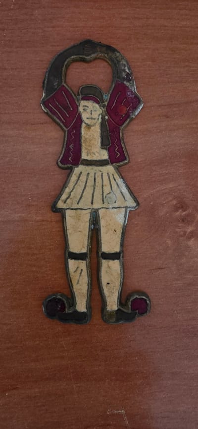 The image shows a piece of decorative metalwork on a wooden surface. The metalwork depicts a stylized human figure in a marching or dancing pose, oriented horizontally. The figure has a pale face with simple facial features. It wears a maroon jacket with a zigzag trim and a light-colored kilt or skirt with vertical lines. The figure also has light-colored legs with maroon accents on the feet and knees, possibly representing boots or stockings. The metalwork appears to be old and shows signs of wear. The wooden surface has a reddish-brown, grained texture, consistent with a piece of furniture or a wall. No specific activity or event is taking place, as the image is a still shot of the object. No text is visible in the image. The location context of Huesca, Spain, does not provide any specific visual cues within the image itself.