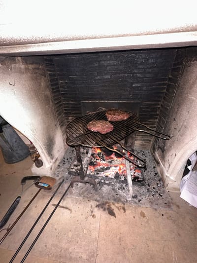 Inside a stone fireplace, two seasoned steaks sit on a metal grill above burning wood. The hearth contains ash and soot, while tools rest to the right, including pokers and a brush. The fireplace interior is lined with dark bricks, contrasting with the lighter stone facade.