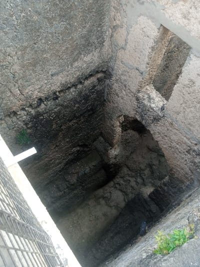 An aerial-perspective image depicts the interior of a deep, square-shaped stone or concrete well or cistern located within the Makadara/Mji Wa Kale ward of Mombasa, Kenya. The rough, textured walls vary in shades of grey and brown, displaying extensive dark staining, consistent with prolonged exposure to moisture and grime, particularly on the upper and mid-level sections. The internal structure features multiple irregular ledges and recesses. At least four dark-plumaged pigeons are present: two are perched on an upper-left ledge, one rests on an L-shaped concrete beam in the upper-right quadrant, and another is situated on a deeper ledge towards the middle-right. Small patches of green vegetation grow from various crevices on the well's walls, notably in the lower-left and lower-right areas. A portion of a light-colored metal grate or cover is partially visible at an upward angle in the bottom-left corner, indicating the well's top opening.