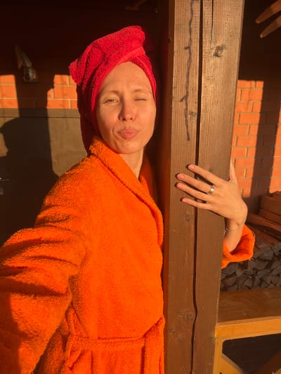 A person wearing an orange bathrobe and a red towel turban poses for a selfie against a wooden post. The background includes a brick wall, a wooden structure, and a stack of firewood. The scene is lit by the warm glow of the sun, creating strong shadows. The image appears to be taken outdoors, possibly in the city of Bol'shaya Shishovka, Russia.