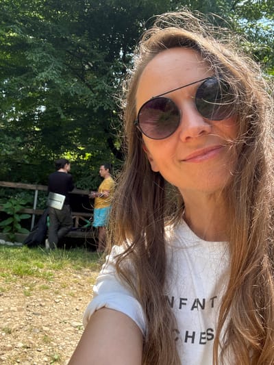 A selfie is taken outdoors, with the subject's face and upper body filling most of the frame. The person has long, light brown hair that is somewhat windswept and falls around their face. They are wearing round, dark-lensed sunglasses. Their expression is relaxed, with a slight smile. They are wearing a white t-shirt with black text that reads "ENFANT TERRIBLES".
In the background, slightly out of focus, two other individuals are visible. One person is wearing a yellow shirt and blue shorts, and the other is wearing a black long-sleeved shirt and dark pants. They appear to be standing near a wooden structure that could be a fence or part of a seating area. The setting is lush and green, with trees and foliage visible behind the people. The ground in the foreground is a mix of dry grass and gravel. The lighting suggests it is daytime, likely sunny given the shadows. The overall mood appears casual and outdoorsy.