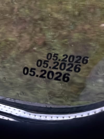 The image shows three lines of text printed on a reflective surface, likely glass, in the city of Topily, Russia. The text reads "05.2026" and is printed in black, bold, sans-serif font. The lines are stacked vertically, with the topmost line slightly offset to the right. The background is blurred and appears to be an outdoor scene with foliage. A portion of a dark, curved object, possibly a window frame or rim, is visible at the bottom of the image, with a series of small circular indentations along its edge. The overall lighting suggests an overcast day.