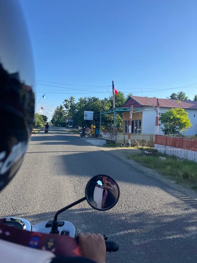 This is a first-person perspective image taken from a motorcycle in Welado, Indonesia. The rider, wearing a helmet, is looking forward down a paved road with vegetation on either side. In the foreground, the motorcycle's handlebars and a portion of the seat are visible. A side mirror reflects the rider's hand and a person holding a phone. In the distance, two flags are flying on a flagpole: one is blue and white, and the other is red and white. A building with a red roof and a yellow facade is situated on the right side of the road, bordered by a white and red striped fence. Power lines are strung overhead. The sky is clear and blue, indicating a daytime scene.