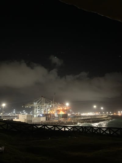 The image captures a nighttime view of a large, illuminated industrial port, identifiable as Tanger Med Port, located near Ksar El Majaz, Morocco. In the immediate foreground, a dark grassy embankment is present, partially obscured by a dark wooden fence. Beyond the fence, a breakwater constructed of numerous concrete tetrapods extends into the water, where waves are observed. The midground and background are dominated by the brightly lit port terminal, featuring multiple large gantry cranes positioned over a quay. At least one container ship is docked, partially visible near the cranes, with shipping containers visible on the vessel and within the terminal area. Various industrial buildings and numerous tall light poles providing extensive artificial illumination are distributed across the port. The sky above is dark, with patches of clouds reflecting the port's lights, and several faint, distant light sources, including a distinct green light, are discernible in the upper dark sky. No individuals are visible. A dark, indistinct overhead element partially frames the top edge of the image.