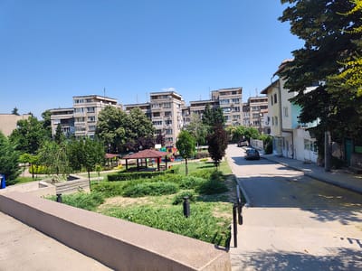 This image depicts an outdoor scene in the city of Svishtov, Bulgaria, likely a residential area, on a clear, sunny day. In the foreground, a concrete barrier with a textured surface runs diagonally across the frame from bottom left to mid-right. Beyond this barrier, a small park area is visible, featuring well-maintained green grass, low bushes, and some scattered small plants. Two black modern-style lampposts are positioned within the grassy area. Further into the scene, a covered wooden gazebo with a red roof stands centrally in the park, and adjacent to it, a colorful children's playground with various structures is present.

Behind the park, a row of multi-story apartment buildings, typical of socialist-era architecture, dominates the background. These buildings are constructed from light-colored concrete blocks and feature numerous windows and balconies. Trees with lush green foliage are interspersed among the buildings and within the park, providing shade. To the right of the apartment buildings, a two-story residential building with a light-colored facade and windows is visible. A paved road runs alongside the park and buildings, with two cars parked on it. The sky is a clear, bright blue, suggesting fair weather. The overall impression is that of a well-developed urban green space within a residential neighborhood.