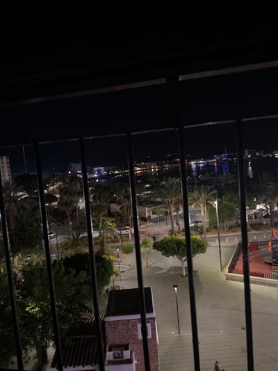 The image shows a nighttime view through horizontal bars, likely a balcony or window. The scene is a waterfront area in Sant Antoni De Portmany, Spain. On the left, a body of water reflects the lights of buildings and boats, suggesting a harbor or marina. The lights create a vibrant, somewhat hazy glow on the water.

In the mid-ground and right, there is a street or promenade lined with palm trees and other vegetation. The ground appears to be paved with a textured material, possibly cobblestones or patterned tiles. Several streetlights are visible, casting pools of light. Buildings with stone and tile facades are also present, with one featuring an outdoor air conditioning unit. A glimpse of a playground structure with orange and blue elements can be seen in the upper right.

The time of day is clearly night, indicated by the darkness and artificial lighting. The weather appears calm, with no signs of rain or strong winds. There is a solitary figure visible on the paved area on the right side of the image, walking away from the camera. The overall mood is peaceful, with the lights on the water and the quiet street creating a serene ambiance. No visible text is present in the image.