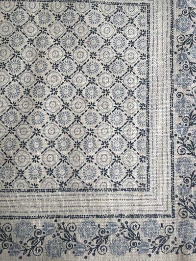 The image depicts a close-up of fabric, possibly a textile sample or a finished product like a cushion or tablecloth. The fabric has a repeating pattern featuring a combination of geometric and floral motifs. The main area of the fabric displays a grid-like design with small, dark star-shaped figures intersecting with diagonal lines. Within each square formed by the grid, there is a light grey circular design resembling a stylized flower. Around the perimeter of this patterned area, there are borders with different designs. One border features a floral pattern with stylized roses and swirling leaf-like details. Another border is a plain strip with a subtle woven texture. The colors are muted, primarily consisting of shades of light grey and dark blue/grey. The fabric appears to be made of a textured material, possibly linen or cotton. There is no visible indication of the image's location within the city of Guildford, United Kingdom.