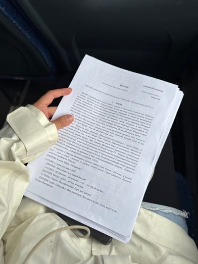 Here's a description of the image:

A hand holds several sheets of printed text. The text is in Russian, and appears to be a script or story titled "Kosmos" by Aleksei Zhitkovsky. 

The hand is wearing a white jacket with velcro closure. The person is likely sitting inside a car. Parts of the car's interior, including the seat and trim, are visible in the background.  

The page number "1" is printed at the bottom right corner of the visible page.

There is no visible indication that the image was taken in the city of Morshanskoye, Russia.