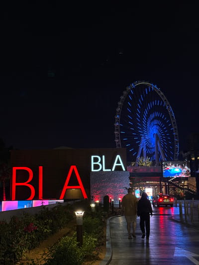 Night scene featuring the illuminated Ain Dubai Ferris wheel. To the right, figures walk on a wet surface reflecting red lights. In the foreground, a sign reading "BLA BLA" is visible, with "BLA" in white and "BLA" below in red. The scene also includes greenery and small path lighting. The overall tone is vibrant and modern.