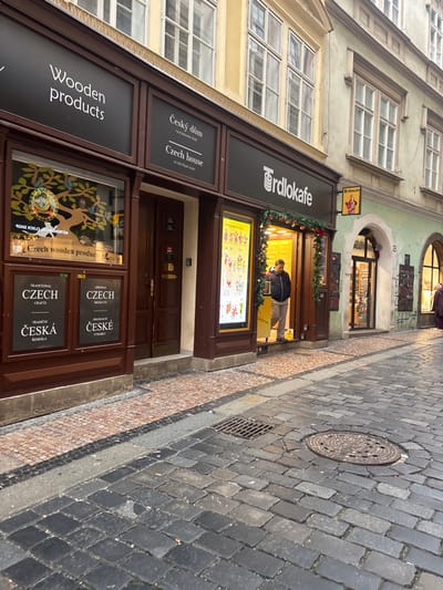 The image depicts a narrow, historic cobblestone street in Capital City of Prague, Czechia, specifically on what is identified as the "Royal route." The scene features the ground-level facades of two multi-story buildings and a segment of the street.

The prominent building on the left and center has a light yellow upper facade with multiple white-framed windows. Its ground floor is clad in dark brown wood paneling, housing several retail establishments. From left to right, a shop displaying "Wooden products" is visible, with its window showcasing a circular clock-like motif on a tree and text reading "Czech wooden product." Below this window are signs in both Czech and English for "Traditional Czech Crafts" and "Original Czech Products." To its right is a dark brown double door. Above these shops, signage reads "Český dům / Czech house on the Royal route." Further right is the brightly lit entrance of "Trdlokafe," a cafe, adorned with green festive garlands, possibly Christmas decorations. A vertical digital display board showcasing various beverages is positioned to the left of the Trdlokafe entrance.

Adjacent to the Trdlokafe, on the right side of the image, is a building with a light green facade and an arched doorway leading to another shop. A small yellow "Trdlokafe" sign is affixed to this building above its entrance.

Two individuals are present:
1.  A male subject wearing a dark blue puffy jacket and light-colored pants stands inside the brightly lit entrance of the "Trdlokafe" shop, facing partially towards the street.
2.  A second person, clad in a dark coat, is partially visible on the far right edge of the image, walking away from the viewer towards the right, near the entrance of the light green building.

The street is paved with dark grey, irregular cobblestones, showing signs of wear. A round metal manhole cover is situated in the street slightly right of center, and a rectangular metal grate (drain) is closer to the left pavement. The pavement directly in front of the shops is composed of smaller, reddish-brown and grey mosaic-like cobblestones, bordered by a dark grey strip that separates it from the main street.
