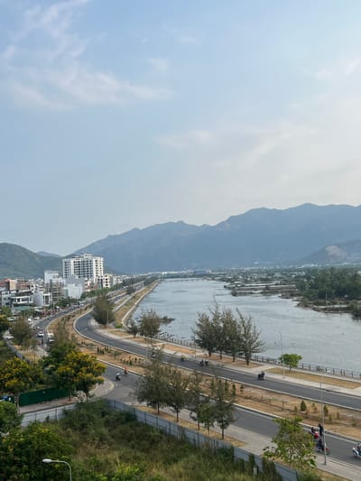 This high-angle image provides a panoramic view of a coastal or riverine landscape, likely in the Nam Natural Heritage Area of Trang, Vietnam. The scene is dominated by a majestic mountain range, a wide body of water, a winding road, and a cluster of buildings, all under a hazy sky.
In the mid-ground, a multi-lane road, possibly a coastal highway, curves and stretches parallel to the water. It features a central divider and is lined with trees and streetlights. Several motorbikes are visible on the road, indicating active daily traffic, with people riding them. To the right of the road, particularly in the foreground, dense green foliage, including trees with some yellow blossoms, fills the space.
To the left of the road lies a broad body of water, appearing calm and somewhat murky, possibly a river or a bay. Along its far bank or within the water, some structures or small vessels can be seen. Further across the water, a collection of modern, white multi-story buildings forms an urban or residential area nestled at the base of the mountains.
The background is dominated by a vast range of mountains, their slopes covered in lush, dark green vegetation. These mountains extend into the distant, hazy horizon, providing a dramatic backdrop to the scene. The sky above is a pale, hazy blue with subtle, indistinct white cloud formations, suggesting either an overcast day or atmospheric haze.
The image captures a daytime scene, characterized by diffused natural light. The overall impression is one of a busy yet scenic coastal environment, with the natural beauty of the mountains and water contrasting with the human development along the shore. No visible text is discernible in the image.