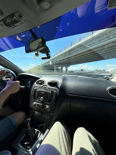The image shows the interior of a car, viewed from the passenger seat, with a driver's hands on the steering wheel. The car is on a highway, passing under an elevated road. The scene appears to be in Novosaratovka, Russia, given the context provided. The driver is wearing a blue shorts and the passenger is wearing light gray sweatpants. The dashboard features a Sony audio system, air vents, and a gear shift. A rearview mirror with a dashboard camera and an air freshener hangs from the windshield. The sky is blue with some clouds, and sunlight is casting shadows. The elevated road has guardrails and supports, and the surrounding landscape includes grassy slopes and other vehicles on the highway.