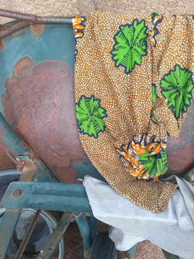 The image shows a close-up view of a portion of a rusty, teal-colored wheelbarrow in Zinna, Nigeria. A colorful fabric with a pattern of green floral designs on an orange and white dotted background is draped over the wheelbarrow's basin. A light grey garment is also visible beneath the fabric. The ground appears to be a mixture of dirt and gravel. The image is taken from a high angle, focusing on the texture and detail of the wheelbarrow and the fabric.