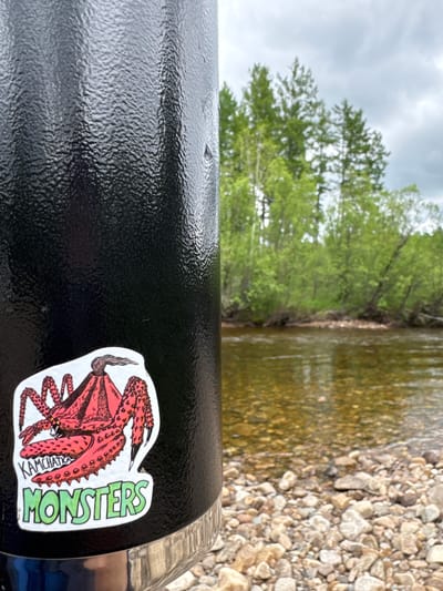 An extreme close-up shot focuses on a black, textured thermos with a sticker of a red crab-like monster and the words "KAMCHATKA MONSTERS" on it. The background is blurred, showing a shallow, clear river with a rocky bed, surrounded by lush green trees and a cloudy sky. The scene appears to be set in a natural environment, possibly near a riverbank. Given the sticker's text, the location might be related to Kamchatka, a peninsula in the Russian Far East. The specific location within the city of Murtygit, Russia, cannot be determined from the image alone.