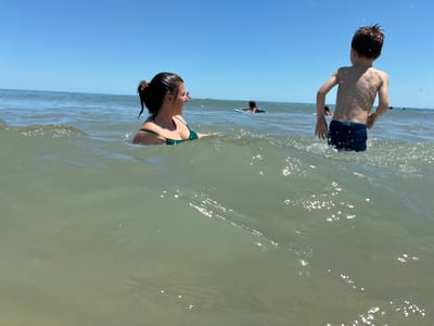 A medium shot captures a vibrant summer scene at the beach in Shkorpilovtsi, Bulgaria. The foreground is dominated by the greenish-blue water of the Black Sea, with a small wave cresting in the center of the frame. To the left, a young woman with dark hair pulled into a ponytail, wearing a green bikini top, is partially submerged in the water, looking to her right with a joyful expression. Her mouth is open, suggesting she is laughing or speaking. In the background, a young boy with short brown hair, wearing blue swim trunks, stands in the water, his back to the camera. He is splashing and appears to be engaged in play. Further in the background, other beachgoers can be seen in the water, one of whom is on a blue flotation device. The sky above is a clear, bright blue, indicating a sunny day. The overall impression is one of a happy, active summer day at the beach.