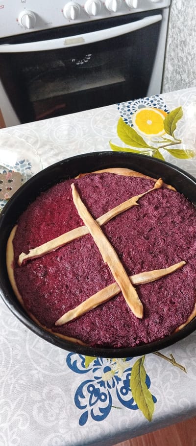 The image shows a close-up, overhead view of a dark purple pie in a black pie pan, placed on a table covered with a tablecloth. The pie has a lattice crust made of light-brown pastry strips arranged in an X shape. The pie filling appears textured and slightly uneven. To the left of the pie pan, a portion of a metal kitchen utensil with a perforated surface is visible. In the background, the lower part of a white oven with black knobs is visible. The tablecloth has a patterned design featuring blue floral motifs and a slice of orange with green leaves. The scene is lit by natural light, casting subtle shadows. The overall setting suggests a domestic environment, likely a kitchen. The image does not provide any specific indicators for the location of Подгорное, Russia.