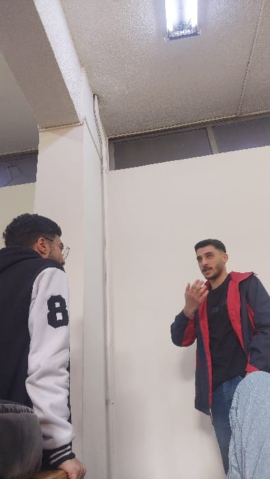 The image depicts an indoor scene within the Al-Qaboun Municipality, Syria, featuring two young adult males engaged in an interaction. The environment consists of white walls and a light-colored, textured ceiling from which a rectangular fluorescent light fixture is suspended.

On the left, a male subject is partially visible from the rear-left perspective. He has dark, curly hair and a beard, wearing glasses. His attire includes a black hoodie with white sleeves, the right sleeve displaying a prominent black number "8". His left arm is resting on a wooden surface, possibly a table or a chair back. He is oriented towards the second subject.

On the right, a second male subject is visible from the chest up, facing partially towards the camera and the first subject. He has short, dark hair and a trimmed beard or stubble. He is wearing a black t-shirt underneath a dark blue jacket with red inner lining or accents. His right hand is raised and slightly open, in a gesture consistent with speaking or explaining. His mouth is slightly open, suggesting active speech. A light blue, possibly textured fabric, is visible in the lower right foreground, partially obscuring the lower body of the second male subject or an unseen third individual.

The interaction suggests a conversation or discussion between the two visible males, with the second male actively gesturing while speaking.