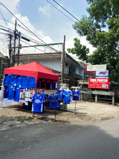 A street vendor selling blue soccer jerseys is set up on the side of a road in the Kecamatan Cimahi Selatan, Indonesia. The vendor has a red canopy tent and a variety of jerseys displayed. A man is sitting in front of the stand. There is a sign for "JNE Express" and a "TOKO PRINTER" sign. The road is paved and there are a few cars in the background. There is a building on the other side of the street with a fence in front of it. The sky is blue with some white clouds. There are many overhead power lines. The scene is in the Kecamatan Cimahi Selatan, Indonesia. The coordinates are -6.909, 107.568.