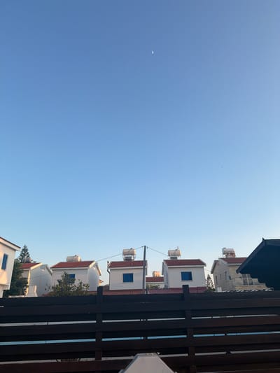 The image captures a view of houses in Pervolia, Cyprus, against a clear, pale blue sky. In the upper part of the sky, a thin crescent moon is visible. Several houses, characterized by their white walls and red-tiled roofs, are lined up in the mid-ground. Some of these houses have cylindrical water heaters on their roofs, and blue-shuttered windows are visible. A dark wooden fence spans the foreground, obscuring the lower portions of the houses. The scene appears to be set during either early morning or late afternoon, given the soft lighting.
