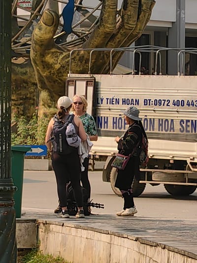 Three individuals are observed conversing on a tiled sidewalk in Sa Pa, Vietnam, under bright daylight. A person on the left, viewed from behind, wears a white baseball cap, dark tank top, black leggings, and athletic shoes, carrying a black backpack with a grey garment attached; trekking poles rest near their feet. Facing them is another individual with blonde hair, sunglasses, and a green and white patterned short-sleeved shirt. To their right, a third person in side profile wears a grey bucket hat, a dark long-sleeved shirt with embroidered details, black pants, and light-colored athletic shoes, carrying a patterned shoulder bag and a dark backpack, holding a small object to their mouth area.
In the background, a large, golden-toned sculpture of a hand is prominent, partially obscured by metal scaffolding or framework. A large white truck is parked behind the individuals, displaying Vietnamese text on its side, including "LÝ TIẾN HOAN ĐT:0972 400 443" and "ANH HỘI ON HOA SEN". A dark green trash bin and a blue sign with a white arrow pointing right are visible on the far left of the frame, near the edge of the paved street. The sidewalk is elevated, with vegetation growing below the curb.