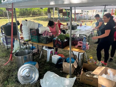 The image captures an outdoor scene under a white tent, likely part of a food service or preparation event, possibly a community gathering or festival. Three women are visible, engaged in activities related to food handling and service.

The woman in the foreground, wearing a black t-shirt and glasses, is looking to the left, her hands in motion, suggesting she is involved in serving or preparing food. To her left, another woman in a grey apron is standing by a table covered with a white cloth, seemingly organizing items or interacting with food. Further left, a woman in a pink shirt is bending over a table laden with various food items and containers, possibly sorting or preparing ingredients.

The scene is set outdoors, with dry grass and vegetation on the ground. In the background, a playground structure with colorful elements is visible, along with trees and a fence. Several tables are set up, some with chairs, indicating a larger event. Various objects are scattered around: large metal pots, plastic buckets, crates filled with what appear to be vegetables like cucumbers and tomatoes, plastic bottles of water and oil, cardboard boxes, and stacks of white disposable containers. A red strap hangs from the tent pole.

The lighting suggests it's daytime, with bright sunlight casting shadows, indicating a warm, possibly summer day. The overall mood appears to be one of busy activity and preparation.

Visible text includes "DEVIN" on a blue plastic water bottle and "FIESTA" on a cardboard box. The text on the black t-shirt of the woman in the foreground is partially obscured but appears to be in a non-Latin script.