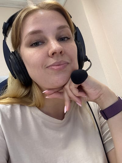 A young woman with blonde hair, wearing a light-colored t-shirt and headphones with a microphone, is captured in a close-up, selfie-style shot. Her left hand rests on her cheek, with manicured pink nails visible. She is looking towards the camera with a neutral expression. The background appears to be an indoor setting with a textured ceiling. A purple smartwatch is visible on her wrist. The image is oriented vertically, as if taken with a phone. There are no visible text elements. The context provided indicates the location is Заволжье, Russia.
