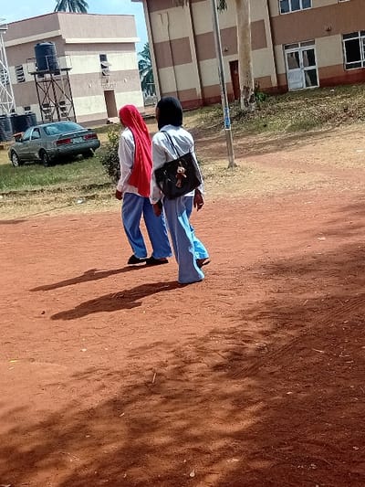 The image displays an outdoor scene featuring two individuals walking on a reddish-brown unpaved ground, possibly a dirt track or pathway. The perspective is slightly from behind and to the side of the individuals, looking towards a series of buildings in the background.
The two individuals appear to be young people, possibly students, dressed in similar attire suggesting a uniform. The person on the right wears a black headscarf, a white long-sleeved shirt, and light blue trousers. They are carrying a black shoulder bag with a white element visible on it. The person on the left wears a bright red headscarf, a white long-sleeved shirt, and light blue trousers. Both individuals are walking forward, casting long shadows to their left on the dusty ground, indicating either morning or late afternoon sunshine.
In the background, there are multi-story buildings with light-colored walls and horizontal brown stripes. Rectangular windows are visible on these buildings. Closer to the foreground on the left side of the image, there is a dark-colored sedan car parked on a grassy patch. Adjacent to the car, some structures resembling elevated water tanks on metal scaffolding are visible. Further in the background, some green vegetation, including what appears to be a palm tree, can be seen. A tall, light-colored pole, possibly a street light or utility pole, stands vertically in the middle distance between the walking individuals and the main buildings. The sky above is clear and light blue.
The overall impression is a sunny day at what might be a school or college campus, with students moving between buildings. No legible text is visible in the image.