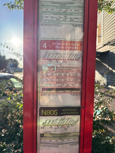 The image shows a red bus stop sign in Getafe, Spain. The sign displays bus route information, including schedules and destinations. One prominent route is line 4, which travels between Perales del Río and Hospital de Getafe. Another route shown is line N805, connecting Madrid (Atocha) to Getafe (Los Molinos). The sign also provides contact information for Avanza Movilidad Integral, S.L., the bus operator. The bus stop appears to be located outdoors, with sunlight filtering through trees and foliage in the background. There are no people visible in the image.