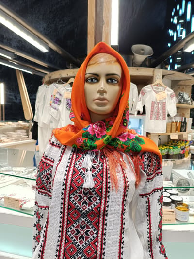 The image shows a mannequin wearing a traditional Romanian blouse and an orange headscarf with floral embroidery. The mannequin is displayed in a market or shop setting, with various items for sale in the background. These include other traditional blouses on hangers, bottles of beverages, and jars of products. A price tag reading "IE TRADIȚIONALĂ COPII" (traditional blouse children) and "Preț: 108,0 lei" is visible. Another price tag reads "43,0 lei" and indicates a perfumed product. The setting suggests a tourist-oriented shop, likely in the city of Turda, Romania, given the Romanian currency (lei) and the nature of the traditional clothing. The lighting appears to be artificial, with fluorescent lights visible overhead.