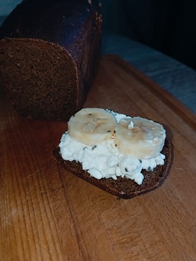 The image shows a slice of dark bread topped with cottage cheese and sliced banana, placed on a wooden cutting board. Part of the loaf of dark bread is visible next to the slice. The location information, Yurgamysh, Russia, is outside the scope of the visible content and cannot be verified from the image.