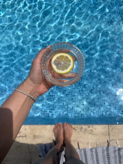 A first-person perspective shot shows a hand holding a glass of water with a lemon slice, over a sun-dappled swimming pool. The hand, adorned with a simple braided bracelet, is positioned towards the center of the frame, with the glass of clear water and lemon taking up a significant portion of the upper half. The pool's turquoise water, with visible tile patterns beneath, reflects the sunlight creating a shimmering effect. The foreground features the edge of the pool, a light-colored tiled border, and a towel on which bare feet are resting. The scene suggests a leisurely moment, possibly in a residential area or a hotel, given the presence of a swimming pool. The location is specified as Beşiktaş, Turkey, which is a vibrant district in Istanbul known for its lively atmosphere, historic sites, and coastal areas, implying this scene could be part of a private residence or a hotel with a pool in that area.