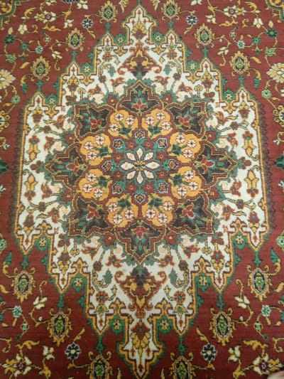 The media file is a close-up, overhead shot of a traditional Uzbek carpet, likely made in Unkurgan, Uzbekistan. The carpet features a central medallion design with intricate geometric and floral patterns. The dominant colors are deep red, cream, yellow, and green, with accents of dark brown and black. The central medallion is surrounded by a series of smaller decorative elements and floral motifs, all set against the rich red background of the carpet. The texture of the carpet is visible, indicating it is woven from threads. The lighting appears to be neutral, with no strong shadows, suggesting an indoor setting or an overcast day. There are no people, animals, or any specific activity depicted; the focus is solely on the artistry and detail of the carpet. The image serves as a visual representation of a handcrafted textile, highlighting its complex design and vibrant color palette, characteristic of Uzbek rug-making traditions.