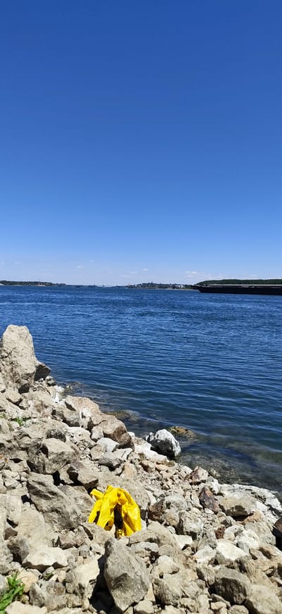 The image shows a rocky shoreline with a bright yellow plastic bag caught among the stones. Beyond the shoreline is a wide, calm body of blue water, likely the Danube River, with a distant cityscape and bridge visible on the horizon. The sky is a clear, vibrant blue. The scene suggests a somewhat remote or natural area along the Vidin embankment, with the presence of litter indicating human impact on the environment.