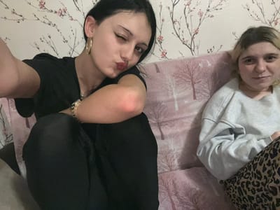 Two females are depicted seated on a sofa within an indoor residential setting. The specific city of Lanchkhuti, Georgia, cannot be determined from the visual information presented.
The female on the left has dark hair, is wearing a black short-sleeved top and black leggings. She wears gold hoop earrings and multiple gold-colored bracelets on her left wrist. Her right eye is closed in a wink, and her lips are pursed. Her left arm is extended forward, holding the camera, indicating she is taking a self-portrait. Her right arm is bent at the elbow.
The female on the right has light-colored hair and is wearing a light gray long-sleeved sweatshirt and leopard print pants. She is seated next to the first female, looking directly towards the camera with a neutral expression.
The sofa is covered with a light pink fabric featuring a repeating pattern of stylized bare trees in white and brown/pink. The wall behind the subjects is covered with wallpaper patterned with cherry blossom branches, displaying pink flowers and brown stems on a light background. The scene is illuminated by interior artificial lighting.