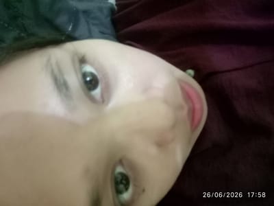 This is a close-up, low-angle, self-taken photograph of a young person lying down, with their face partially obscured by their hand. The subject's eyes are open, and they appear to be looking at the camera. Their skin tone is light. The background consists of dark fabric, possibly bedding or clothing, in shades of dark gray and deep red/maroon. There is a small green object visible on the right side of the frame, near the edge. The lighting is dim, suggesting an indoor setting, possibly at night or in a poorly lit room. The image also contains a timestamp in the bottom right corner: "26/06/2026 17:58". The overall mood of the image is intimate and casual, typical of a selfie taken in a private space. No specific activity or event is clearly depicted, other than the act of taking a photograph of oneself.