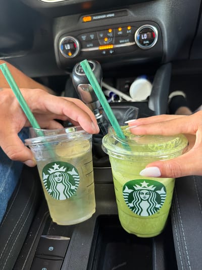 Two Starbucks drinks are held by two individuals in the passenger seats of a car. The drink on the left is a pale yellow, translucent liquid with ice and a green straw. The drink on the right is a bright green, opaque beverage with a green straw. Both cups have the Starbucks logo. The car's interior is visible, including the gear shift, climate controls, and a parking brake button. The scene appears to be in the city of Arroyomolinos, Spain.