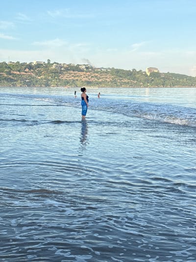 A medium, vertical shot depicts a beach scene. The view is slightly rotated counterclockwise. A woman stands in the shallow water, facing away from the camera, her body angled slightly towards the left. She has dark hair and wears a dark top and patterned blue shorts. The water is clear and ripples around her legs, creating a reflection. Other individuals are visible in the distance, further along the shoreline.
The water transitions from the light blue near the shore to a darker hue further out. Waves are visible in the middle ground. The sky is clear blue with light clouds near the horizon.
In the background, a coastline is lined with trees and buildings. It features a mix of residential and commercial structures, including what appear to be hotels or resorts. Based on the buildings, location, and appearance of the beach, the scene is likely in Jimbaran, Indonesia.