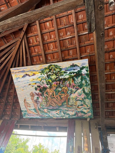 The image shows an overhead view of a tiled roof with exposed wooden beams. A large painting is hung from the beams, depicting a scene with several figures in a lush, tropical setting. The painting features a landscape with trees, water, and distant hills, populated by human figures in what appears to be traditional attire. The style is impressionistic, with bold brushstrokes and vibrant colors. The overall impression is of an artwork displayed in a rustic, possibly outdoor or semi-outdoor environment, common in locations like Tejakula, Indonesia, known for its artistic communities and natural beauty.