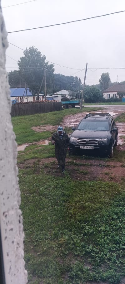 The image shows a rural scene in the village of Podgornoye, Russia, under overcast skies. In the foreground, a person wearing a camouflage jumpsuit and a blue cap is walking on a muddy path. The person is holding something in their right hand. To the right of the person is a dark-colored SUV, likely a Dacia Duster, with a roof rack. The license plate reads "C552PX 18". The ground is wet and muddy, with puddles visible. Behind the SUV, there is a wooden fence separating the yard from a field. Beyond the fence, there are several houses, including one with a blue roof, and trees. A trailer loaded with dark material, possibly hay or manure, is parked near the houses. Power lines and utility poles are visible in the background. The overall impression is of a damp, possibly rainy, day in a rural setting.