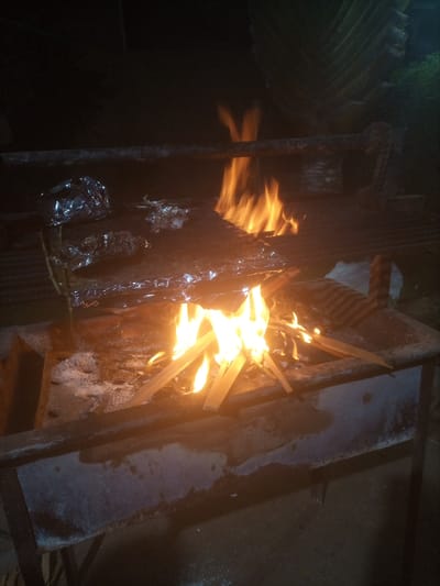 The scene depicts an outdoor nighttime environment in Theta ward, Kenya, illuminated primarily by an active fire. A robust, dark-colored metal brazier or grill occupies the central and lower portions of the frame. Within the brazier's lower section, bright orange and yellow flames emanate from burning wood fragments, arranged in a tepee formation. White ash residue is visible around the base of the fire.

Positioned directly above the flames on a surface, likely a grate, are multiple food items. A large, flat object, entirely encased in reflective aluminum foil, is centrally located. To its left, a smaller, rounded item, also wrapped in foil, rests on the surface. Adjacent to the larger foil package, a small, unwrapped piece of food is discernible.

The metal structure of the brazier shows signs of use and heat exposure, with varied textures and some discoloration. A horizontal metal bar is visible in the upper background, extending across the scene, possibly part of a larger support structure. The background is dark, with a large, fan-shaped leaf structure, resembling a palm frond, visible in the upper right, indicating surrounding vegetation. The ground beneath the brazier appears to be a hard, flat surface, possibly concrete, exhibiting some dampness or reflective qualities. No individuals are present in the image.