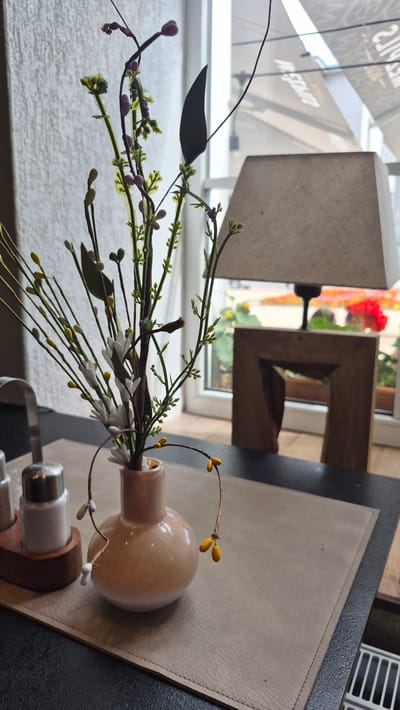 The image shows a close-up, rotating view of a decorative arrangement on a table, likely in a cafe or restaurant. The central focus is a small, light-colored ceramic vase holding an artificial floral arrangement. This arrangement features thin, dark stems with small green leaves and buds, interspersed with tiny white blossoms and yellow teardrop-shaped embellishments. Some branches also have small, pinkish-purple berry-like elements. The vase itself is a pale peach color with a rounded body and a narrow neck.

Behind the vase, the scene suggests an indoor setting with natural light. To the left, a blurred background shows a window with a view of outdoor foliage and what appears to be a street scene, with some signage visible. The signage appears to be in a Latin-based alphabet. A lamp with a textured, light-colored shade is also visible in the background, along with a wooden decorative object with a square cutout.

In the foreground, a light-colored placemat, possibly made of faux leather, is underneath the vase. A salt and pepper shaker set, with a silver top and a dark base, is positioned to the right of the vase. The overall lighting suggests daytime, possibly morning or afternoon, with soft, diffused light. There are no people visible in the image. The scene conveys a sense of calm and decorative ambiance.