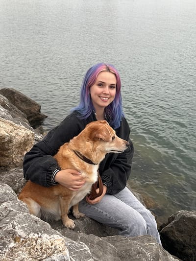 A young adult female with light skin and multi-colored hair (pink and purple) is depicted sitting on large, grey rocks alongside a tan and white medium-sized dog. The scene is set on a rocky shoreline in Gijón, Spain, with a calm, grey-green body of water extending into the background under an overcast sky. The woman, wearing a dark grey oversized jacket with striped cuffs and light blue jeans, is smiling and has her left arm wrapped around the dog. Her right hand is near the dog's chest, holding what appears to be a brown leash or strap. The dog, wearing a patterned dark collar, is seated facing right, away from the camera, between the woman's legs and arm. The rocks in the foreground and midground are rough-textured, varying in size, and display white and brown mineral deposits.