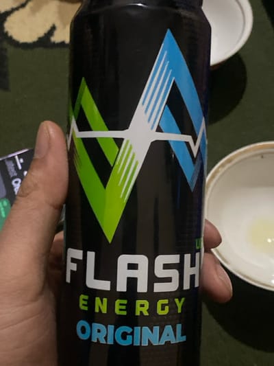 A person's left hand, with a light-to-medium skin tone, grips a black "FLASH ENERGY ORIGINAL" beverage can in a close-up indoor shot in Gulbahor, Uzbekistan. The can's label displays a prominent green and blue stylized lightning bolt or "V" logo above the "FLASH" in white, "ENERGY" in green, and "ORIGINAL" in blue text. In the background, to the left, a portion of a cream-colored patterned rug and a dark electronic device with illuminated green text are partially visible. To the right, two white ceramic bowls are present; the lower bowl features a gold rim and contains a small amount of yellowish liquid. The primary surface beneath the items is dark green.