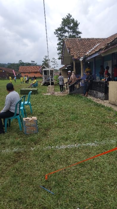 This outdoor photograph, taken in Panjalu, Indonesia, depicts a community gathering or event under an overcast sky during daytime. The image appears to be rotated 90 degrees counter-clockwise.
In the foreground, a person wearing a white long-sleeved shirt and dark pants is seated on a turquoise plastic chair, facing away from the viewer. Another similar chair and a wooden box rest nearby. The ground is covered in green grass, marked with a distinct white painted line and an orange ribbon or tape stretched across it, indicating a designated area for an activity. A blue stake or stick is also visible in the grass.
To the left, a building with a traditional terracotta tiled roof and light-colored walls features a raised porch or patio. Several people, including children and adults, are gathered on this porch. One individual is wearing a light-colored shirt with the number "23" visible on the back. A sound system, comprising speakers on stands, is set up near the building. Further in the background, other buildings with similar architecture and lush green trees are visible. A white power line or wire stretches across the upper part of the frame. The scene suggests a relaxed communal event or celebration in a rural or semi-rural Indonesian setting.