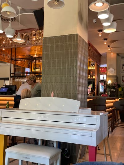 The image displays the interior of a contemporary restaurant, bar, or cafe, illuminated by warm artificial lighting that suggests an evening setting.
In the foreground to the right, a prominent white digital grand piano, branded "YAMAHA," stands with a matching white piano bench also featuring the "YAMAHA" logo. A small pink object rests on the piano's surface.
Behind the piano, a section of a counter or wall is adorned with textured, rectangular gray tiles. Further back, a well-stocked bar area is visible, with shelves holding numerous bottles and glassware, alongside a beverage cooler. Above the bar, a clock hangs on the wall, and a red sign clearly displays the text "CORK LIST."
Two individuals are partially seen behind the tiled counter: an adult with blonde hair wearing a green top, and a child with blonde hair next to them, appearing to be occupied with tasks behind the bar.
The establishment boasts modern white ceiling-mounted light fixtures and wall sconces with exposed bulbs, contributing to the inviting atmosphere. Ornate, possibly gilded, decorative moldings on the ceiling and what appear to be artistic balustrades or railings add a classic touch to the decor. A small security camera is visible on a wall.