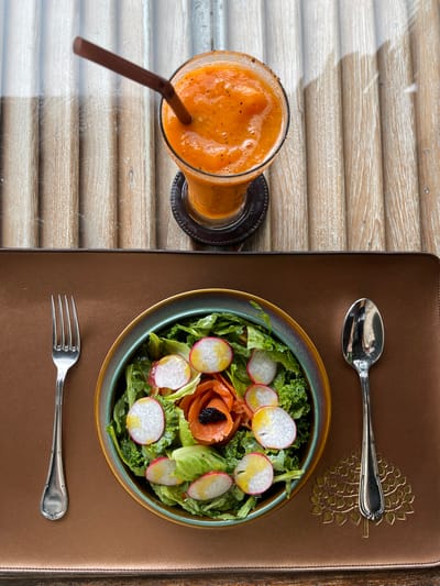 This top-down, overhead shot captures a meal set on a textured, brown placemat beside a light wooden surface. The wooden surface appears to be a table with horizontal planks, possibly in an outdoor or rustic setting, and it is partially covered by what looks like a transparent protective layer, casting faint shadows.
The main subjects are a glass of vibrant orange smoothie and a bowl of salad. The smoothie, in a clear glass, is a bright, pulpy orange color and has a brown straw inserted. It's resting on a dark coaster.
The salad is served in a shallow, multi-toned ceramic bowl with a green rim. It contains a base of leafy greens, topped with thinly sliced radishes that show their pink skins and white interiors, and delicate swirls of smoked salmon. A small dollop of black caviar sits atop the salmon.
A silver spoon is placed above the salad bowl, and a silver fork is positioned below it, suggesting the meal is ready to be eaten. The placemat has an embossed, gold decorative element resembling a stylized tree or foliage in the upper right quadrant.
The lighting suggests daytime, possibly mid-morning or afternoon, given the clear, bright illumination without harsh shadows. The scene evokes a sense of a relaxed meal, likely at a restaurant or cafe, given the presentation and the tableware. The location context of Si Sunthon, Thailand, hints at a warm climate and possibly a beachfront or tropical setting, though these cues are not explicitly visible in the image itself. No text is visible in the image.