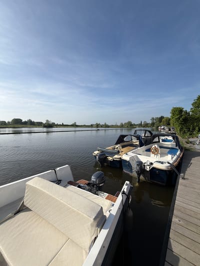 The photograph shows several boats docked alongside a wooden pier, likely in Rhenoy, Netherlands. The scene is captured on a sunny day with a clear blue sky. The water reflects the sky and the opposite shoreline, which is lined with trees. The boats range in size and style, with visible details such as steering wheels, canopies, and outboard motors. Cushions are visible on one of the boats, and ropes tie the boats to the pier.