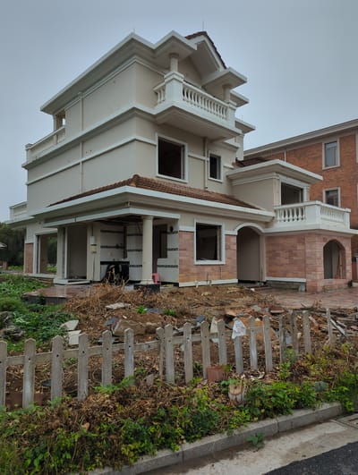 The image depicts an unfinished building in Shunde District, China. The building has a multi-story structure with exposed brick and concrete elements. The exterior lacks windows and doors in certain areas, revealing the interior. Debris and foliage surround the building, and a weathered wooden fence runs along the perimeter. The sky is overcast.