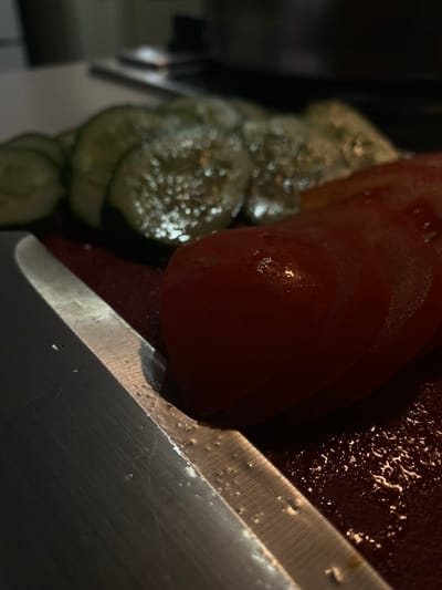 The scene is an indoor setting in Vakhitovo, Russia, depicting a food preparation area. A stainless steel kitchen knife, approximately 20-25 cm in visible blade length, is positioned diagonally across the lower left quadrant of the frame, resting on a dark red cutting surface. The knife blade exhibits multiple small liquid droplets and minor surface scratches. To the right of the knife, several slices of red tomato are arranged, showing glistening moisture on their surfaces. Behind the tomatoes and to the left, multiple slices of green cucumber are visible, also appearing wet. The cutting surface beneath the items is dark red and features scattered liquid droplets, particularly around the knife and vegetables. The scene is dimly lit, with a light source causing specular highlights on the wet surfaces and the knife blade. The background is out of focus and dark. No individuals are present.